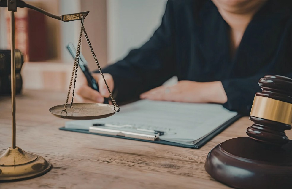 A person filling out paperwork on a clipboard sitting at a desk with a dark wooden gavel and gold scales of justice statue.