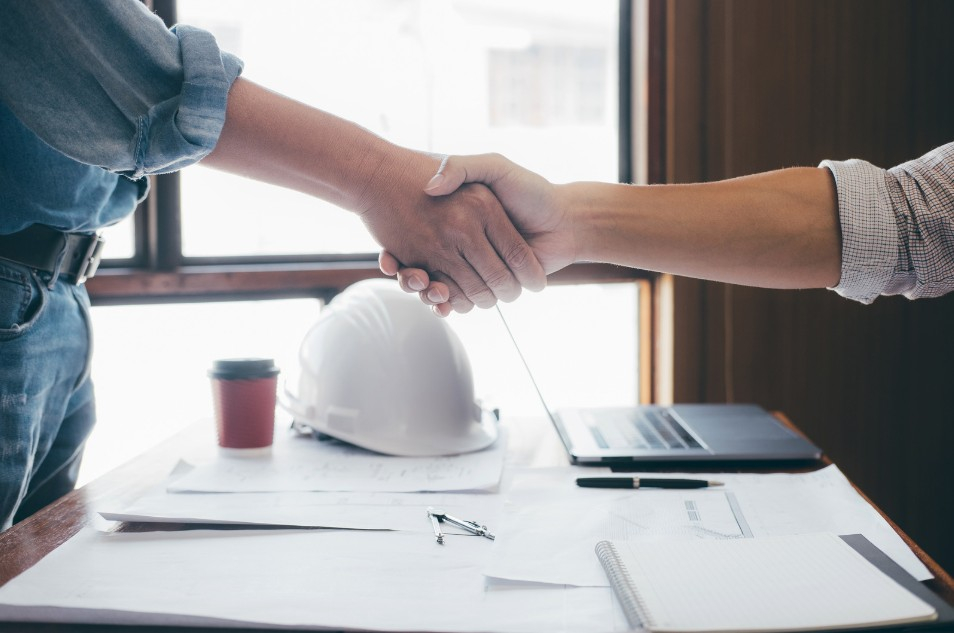 Two people shaking hands above a desk with paper work, a black laptop, and a white hard hat. Two people shaking hands above a desk with paper work, a black laptop, and a white hard hat.