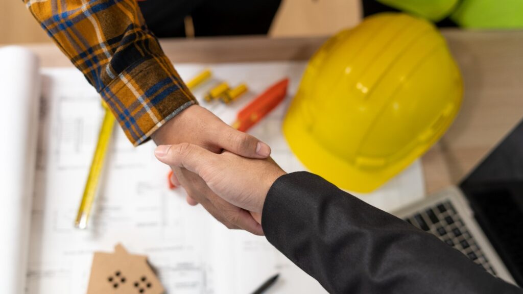 A Close-up of Two People Shaking Hands, in the Background Are House Blueprints.