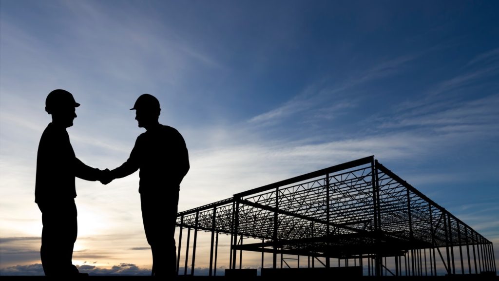A contractor and businessman shake hands in front of a construction site.