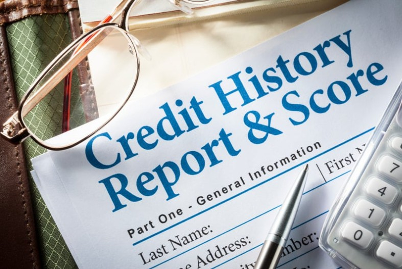 A piece of paper titled “Credit History Report & Score” in blue ink with a silver pen, a white calculator, and oval glasses lying on top.