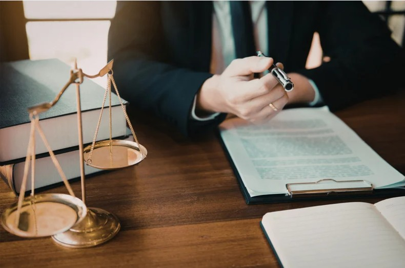 A man in a black suit sits at a wooden desk with a sculpture of the scales of justice holding a pen above a clipboard with paperwork attached to it. A man in a black suit sits at a wooden desk with a sculpture of the scales of justice holding a pen above a clipboard with paperwork attached to it.