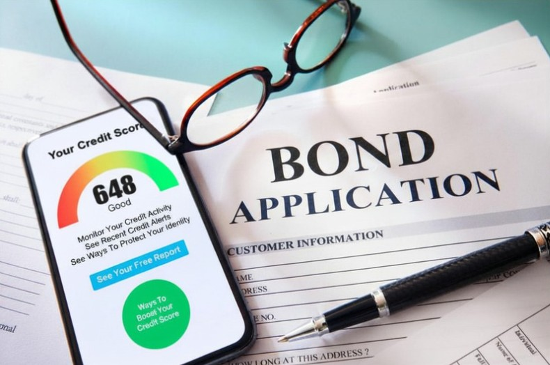A smartphone displaying a good credit score of 648 and a printed bond application form, with a black pen and brown glasses lying on top.