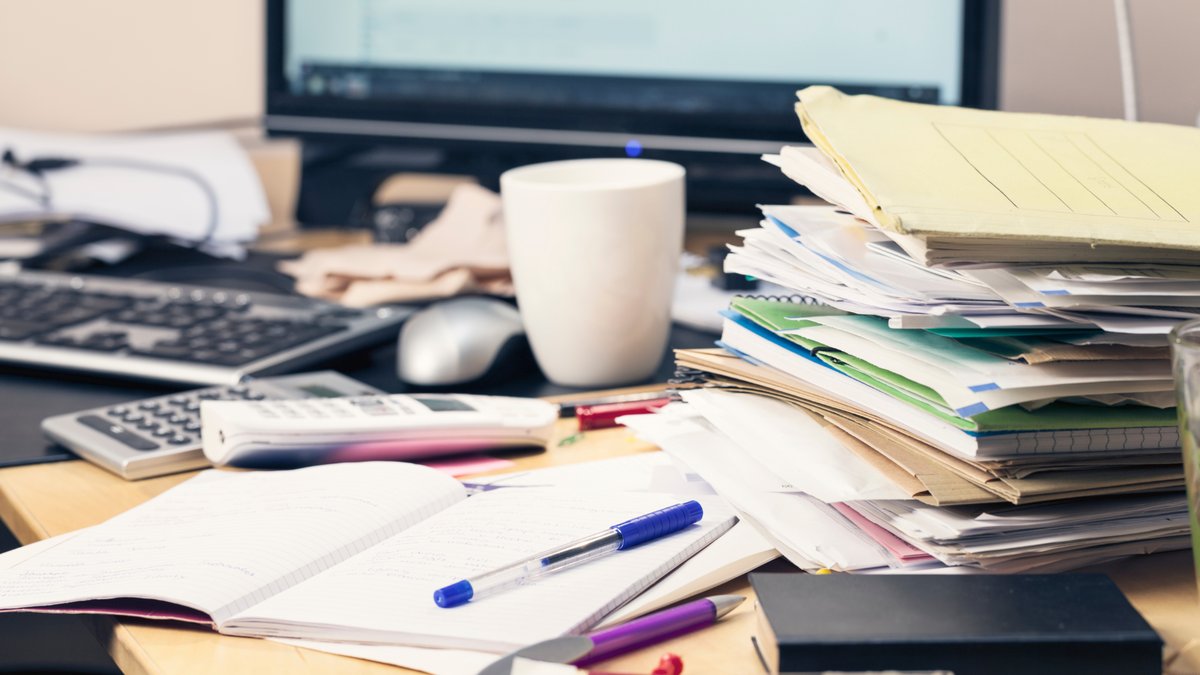 A messy desk covered in file folders, notebooks, pens, and computer equipment